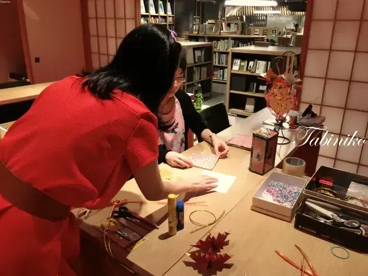一位穿紅裙的女士在擺滿書架的溫馨房間裡，於木桌旁指導客人進行摺紙手工活動。