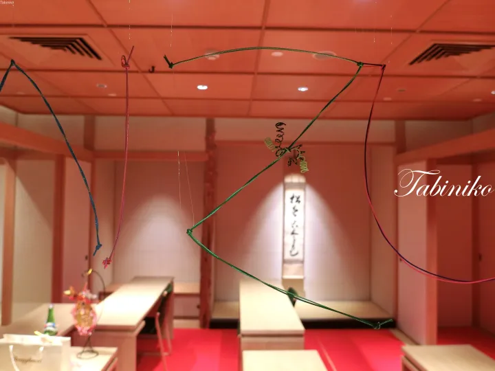 Traditional Japanese dining room with hanging decorative cords and a scroll, featuring the Tabiniko logo.