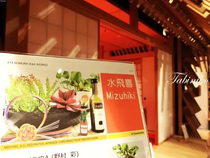 Aya Nomura展出的Mizuhiki展示板，配有植物和酒瓶，背景為溫暖的燈光環境。