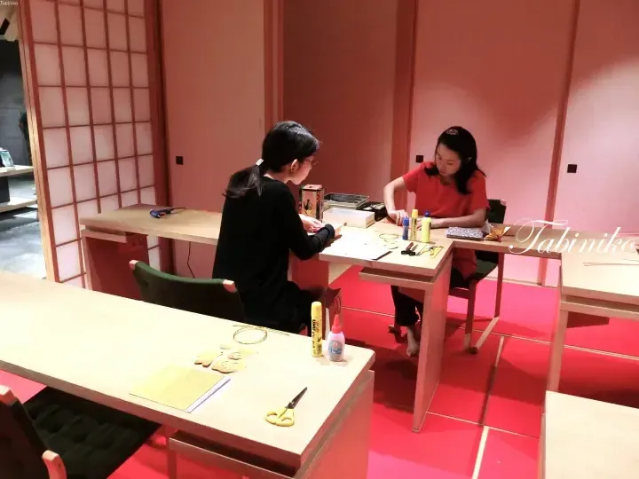 障子のあるピンク色の部屋で木製のテーブルに向かい合う二人の女性。手書きの「Tabiniko」ロゴ入り。