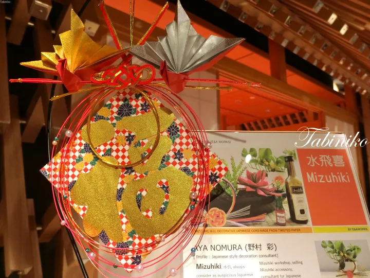 Traditional Japanese Mizuhiki decoration featuring gold and silver paper flowers with a red cord, displayed next to an informational sign.