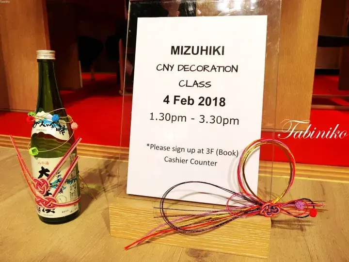 木製テーブルの上に置かれた、色とりどりの紐で飾られた日本酒のボトルとMIZUHIKI CNY装飾クラスのお知らせプレート。