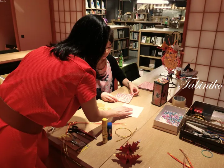A woman in a red dress assists a guest with origami crafts at a wooden table in a cozy, book-lined room.