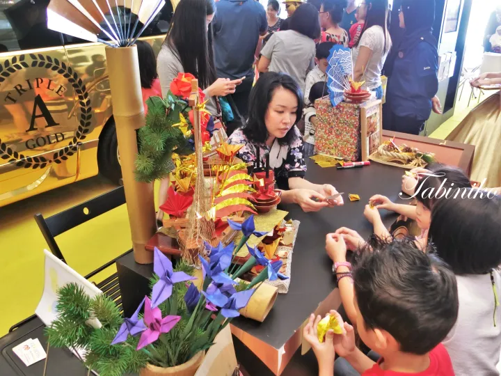 Woman and children making origami at a craft booth with a Triple A Gold award display nearby.