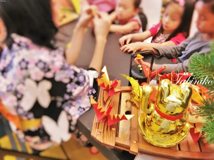 Children gather around a table with a golden Maneki-neko and origami decorations during a cultural activity.