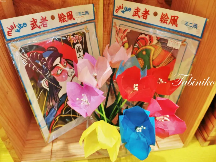Colorful origami flowers displayed in front of packaged mini kite craft kits featuring Japanese samurai designs.