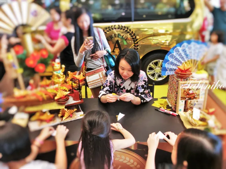 A woman and children fold origami at a table during a golden-themed event with decorative fans and offerings.