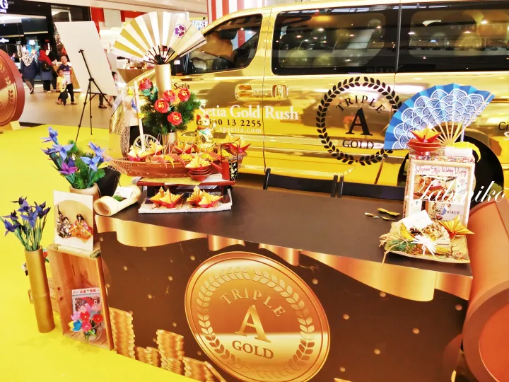 Gold-themed promotional booth featuring a golden van, traditional fans, and decorative displays at a Malaysian event.