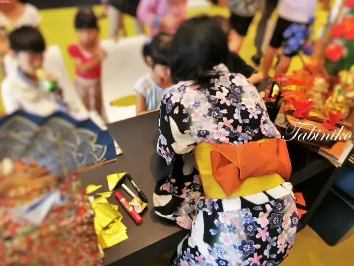 子供たちとテーブルを囲む花柄の着物を着た女性と、色鮮やかな日本の工芸品や装飾品。