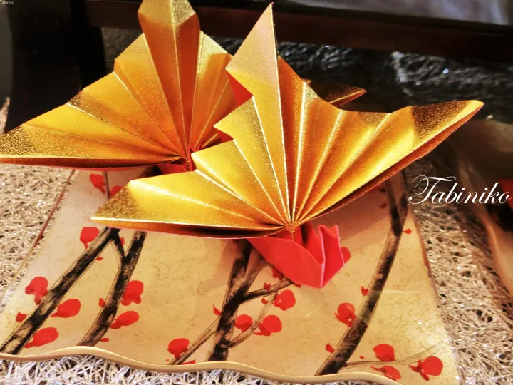 Golden origami crane on a decorative plate with red plum blossoms and a red base.