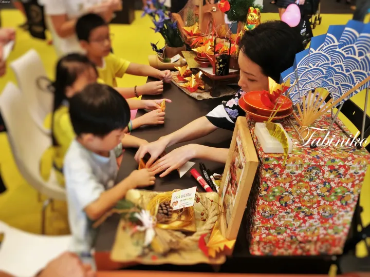 Children and adults participate in a traditional craft activity at a table filled with autumn-themed decorations and materials.