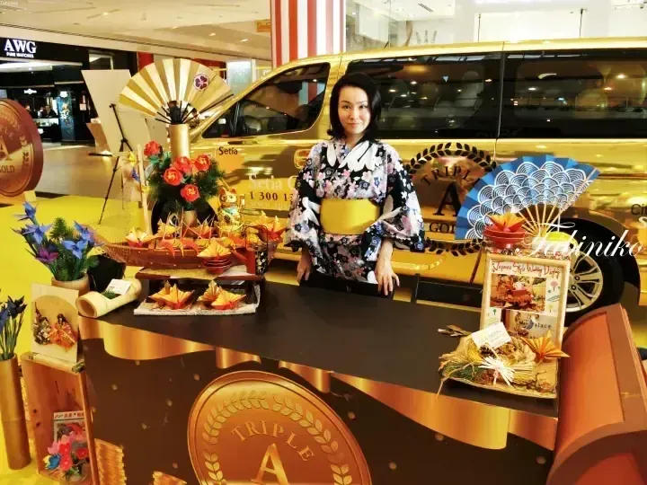 一名身穿花卉和服的女子在購物中心內的Triple A Gold促銷展台前擺姿勢，背景是一輛金色面包車。