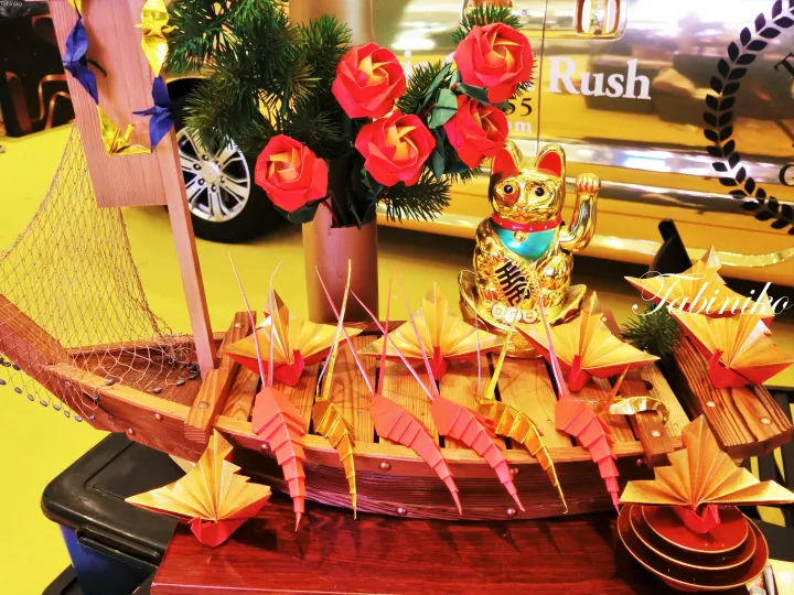 A wooden boat display featuring origami shrimp, a lucky cat figurine, and red roses, with a yellow Rush taxi in the background.