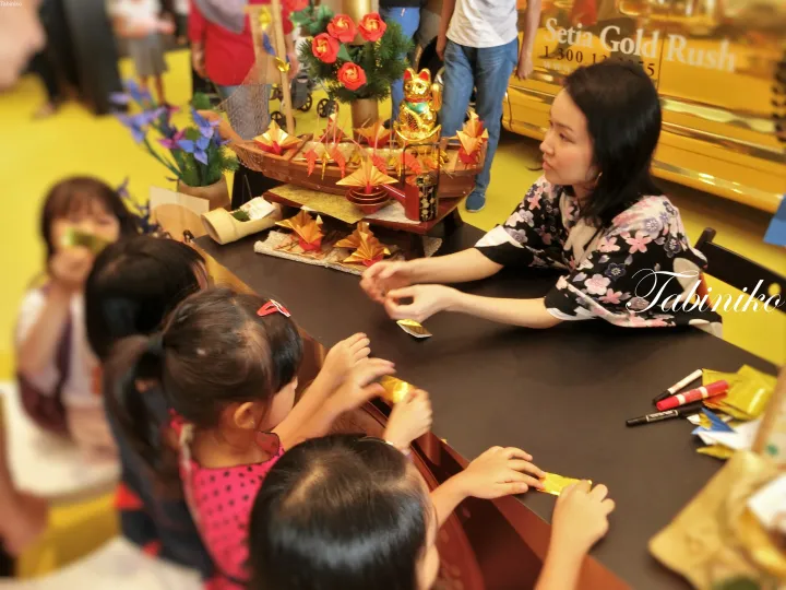 A woman in a floral blouse interacts with children at a table during the Setia Gold Rush event, featuring festive decorations.