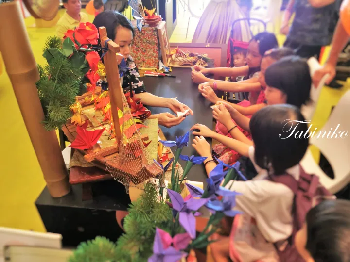 Children learning origami at a cultural display featuring a miniature wooden boat and autumn decorations.