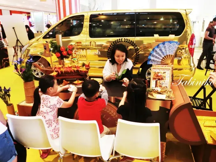 プロモーションイベントでゴールドのホンダ・ゴールドラッシュバン前に座る子供たちに女性が食事を提供している様子。