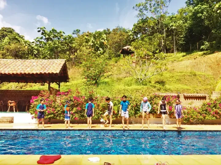Children in swimwear line up at the pool edge at a tropical resort surrounded by lush greenery and bougainvillea.