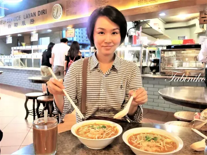 一名女子在自助餐厅吃叻沙，背景有菜单板和其他食客。