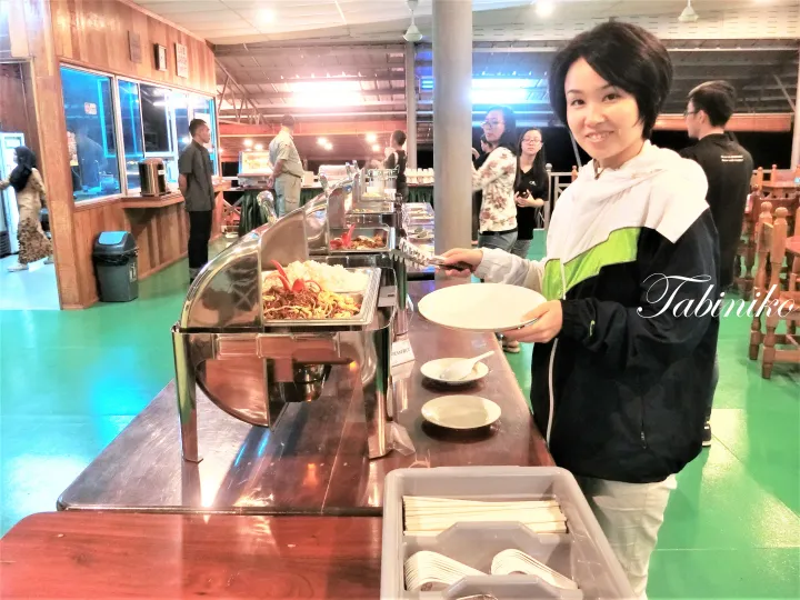 Smiling woman in a black and green jacket holding a plate at a buffet station with stainless steel chafing dishes.