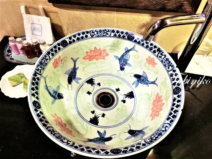 酒店浴室中带有蓝色鱼和莲花图案的装饰性陶瓷洗手盆，旁边摆放着宾客用品