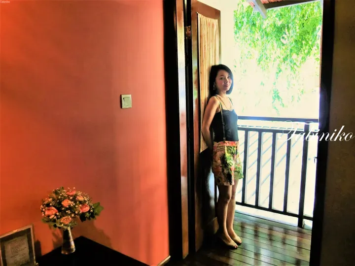A woman stands in a doorway with coral walls, looking out at a balcony with lush greenery.