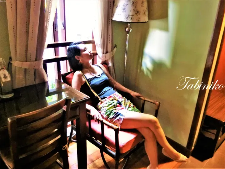 Woman relaxing in a rattan chair at a cafe near a window with curtains and a lamp.