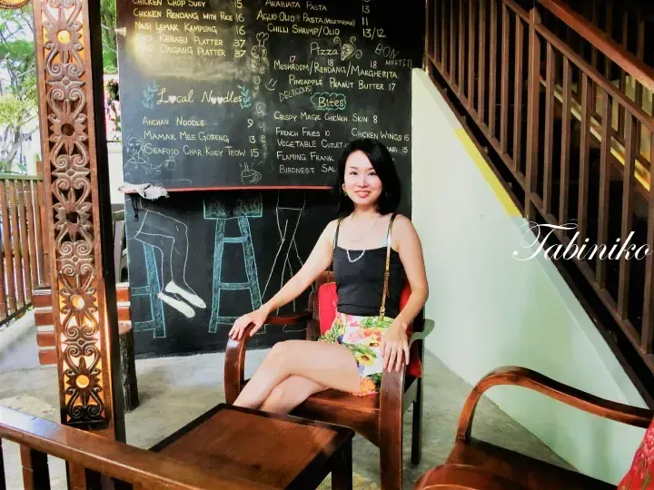 一位女士坐在马来西亚一家咖啡馆的木椅上，背景是写有椰浆饭和当地面条的黑板菜单。