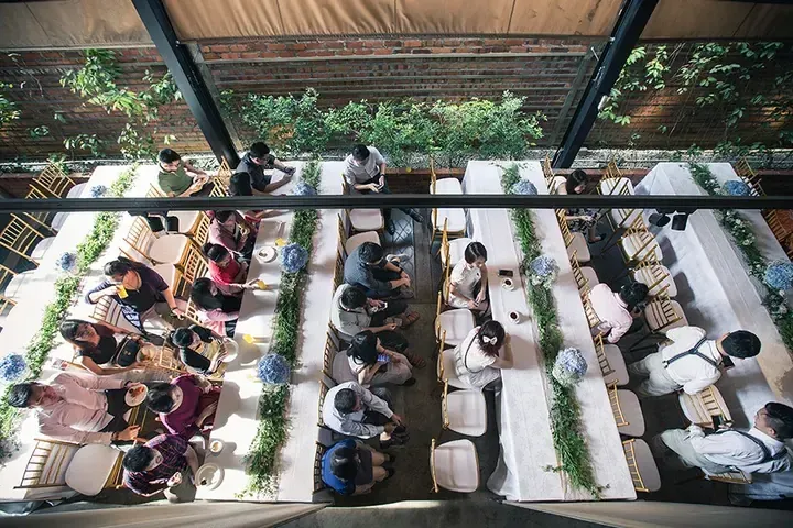 Overhead view of guests dining at long tables in a rustic venue with brick walls and greenery.