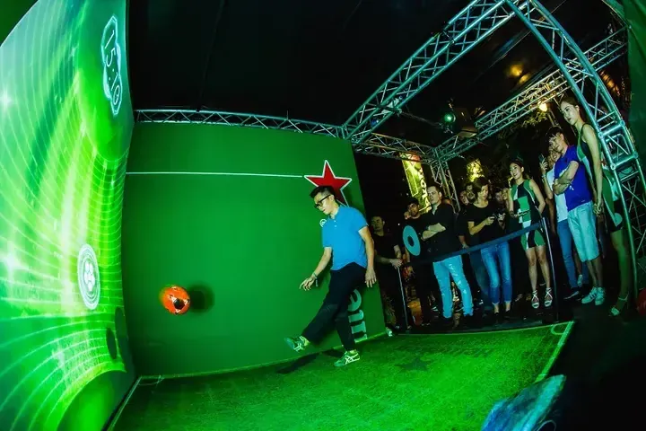 Man kicking a soccer ball at a Heineken interactive green wall display during a lively event.