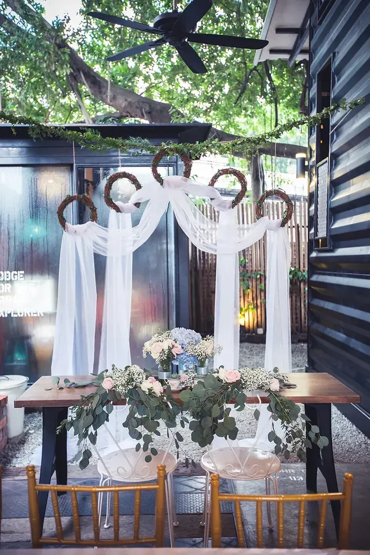 Outdoor wedding setup with rustic wreaths, white drapes, and floral table arrangement at a modern venue.