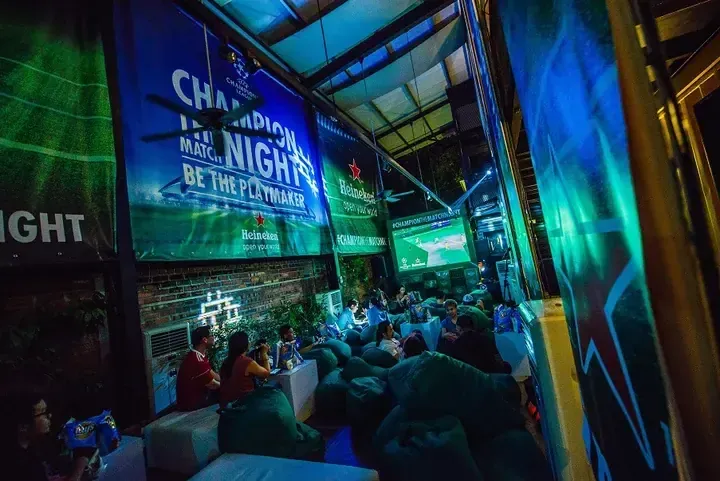 Crowd watches Champions League match on screen at a Heineken event space with bean bags and banners.