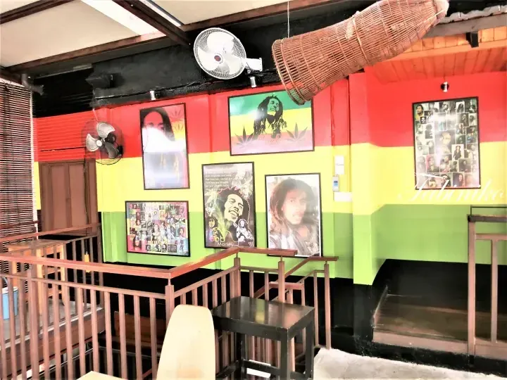 Interior of a Rastafarian-themed cafe with red, yellow, and green walls featuring Bob Marley posters and wooden furniture.