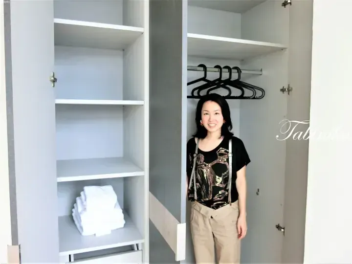 一位面帶微笑的女士站在配有白色層板和掛衣區的現代步入式衣櫃內。