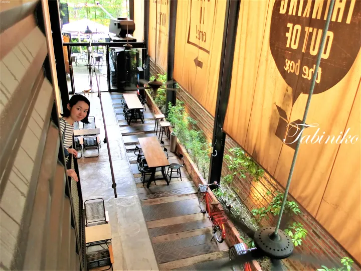 Woman peeking from a balcony overlooking a modern cafe courtyard with wooden tables and a red bicycle.