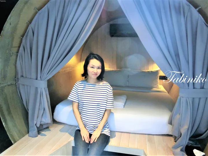 Woman in striped shirt sitting on wooden platform inside a dome-shaped room with blue curtains and a bed.