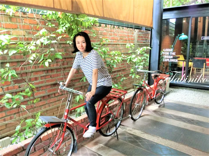 Woman posing with a red bicycle at the entrance of a modern lodge featuring a brick wall and lush greenery.