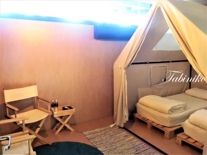 Cozy glamping tent interior featuring a canvas tent bed, wooden director’s chair, and small side table with water bottles.