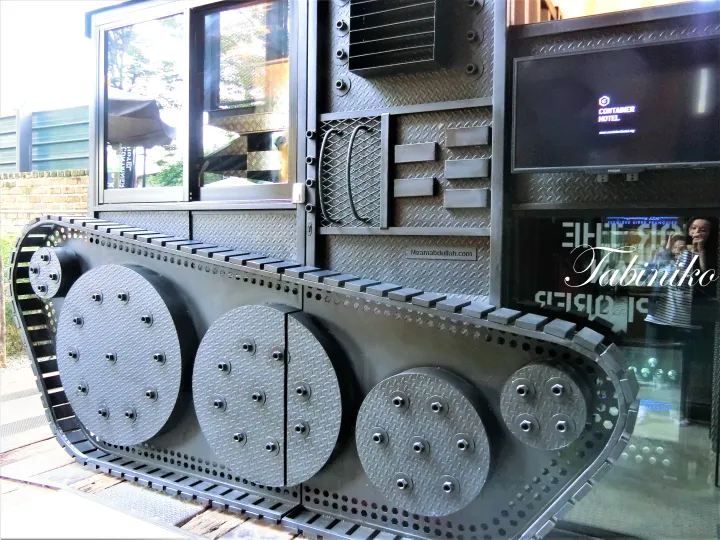 Industrial-style container hotel exterior featuring a large tank tread design and a screen displaying ‘CONTAINER HOTEL’.