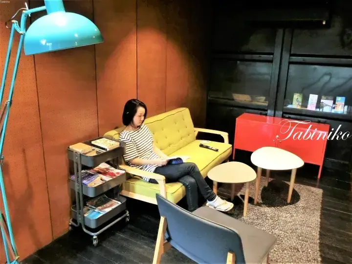 在配有蓝色台灯和书架的舒适休息区，一名女子坐在黄色沙发上阅读书籍。