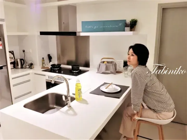 Woman sitting on a stool in a modern white kitchen with a sink, stove, and island counter.