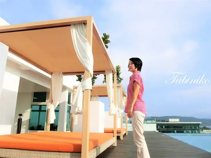 Woman standing on a wooden deck beside orange cushioned daybeds at a resort infinity pool overlooking the sea.