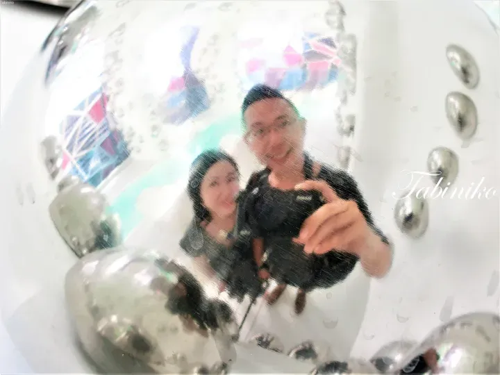 A couple taking a selfie in a distorted reflection on a shiny metallic surface, with the watermark Tabiniko visible.