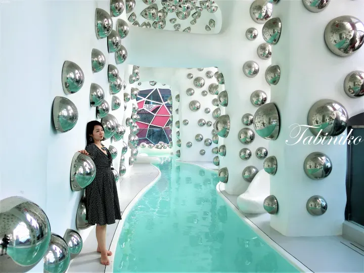 Woman in polka-dot dress posing beside a turquoise pool in a white room decorated with numerous silver dome mirrors.
