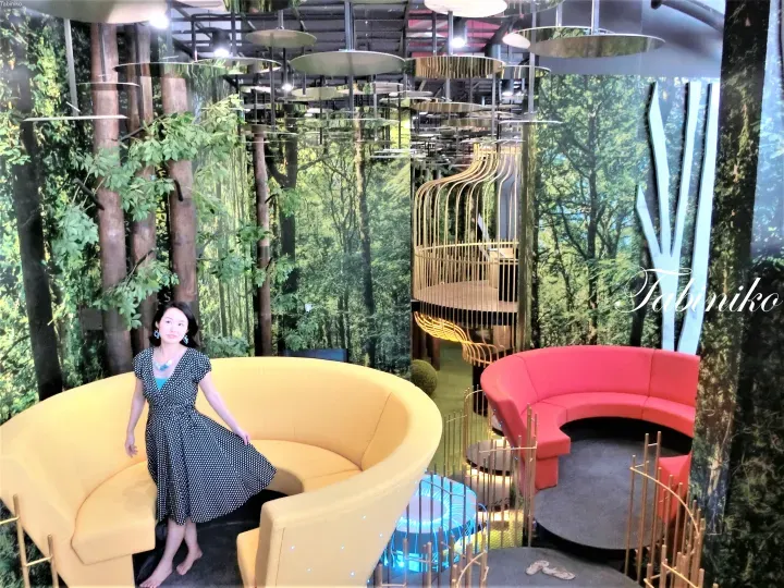 Woman in polka-dot dress posing by a yellow sofa in a modern lounge with forest-themed decor and curved seating.