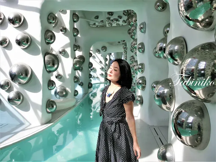 Woman in polka dot dress standing in a white corridor lined with reflective silver spheres and a turquoise pool.