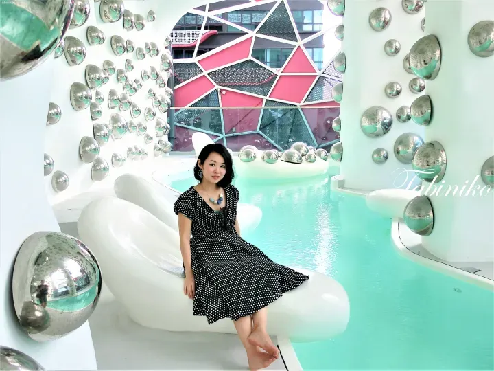 Woman in polka-dot dress sitting on white lounge chair beside pool at W Kuala Lumpur with metallic bubble wall decor