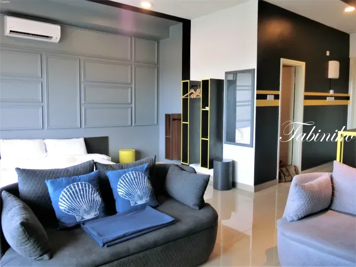 Modern hotel room interior featuring a grey sofa with blue shell pillows, a bed with grey paneled walls, and black shelving with yellow accents.