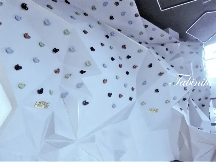 White geometric climbing wall with colorful holds and caution signs in a modern indoor facility.