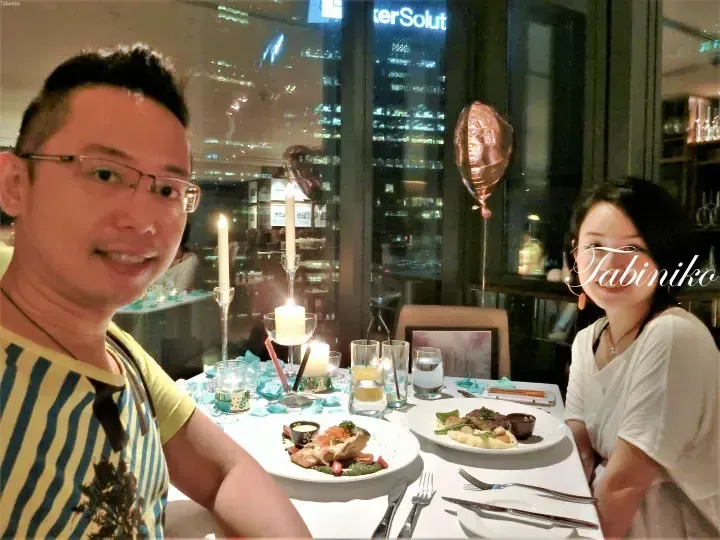 A couple dining at a candlelit table with city views, featuring heart-shaped balloons and plated meals.