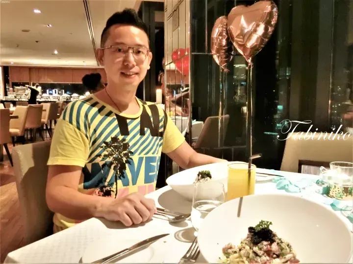 A man smiles at a restaurant table with heart-shaped balloons and plates of food.